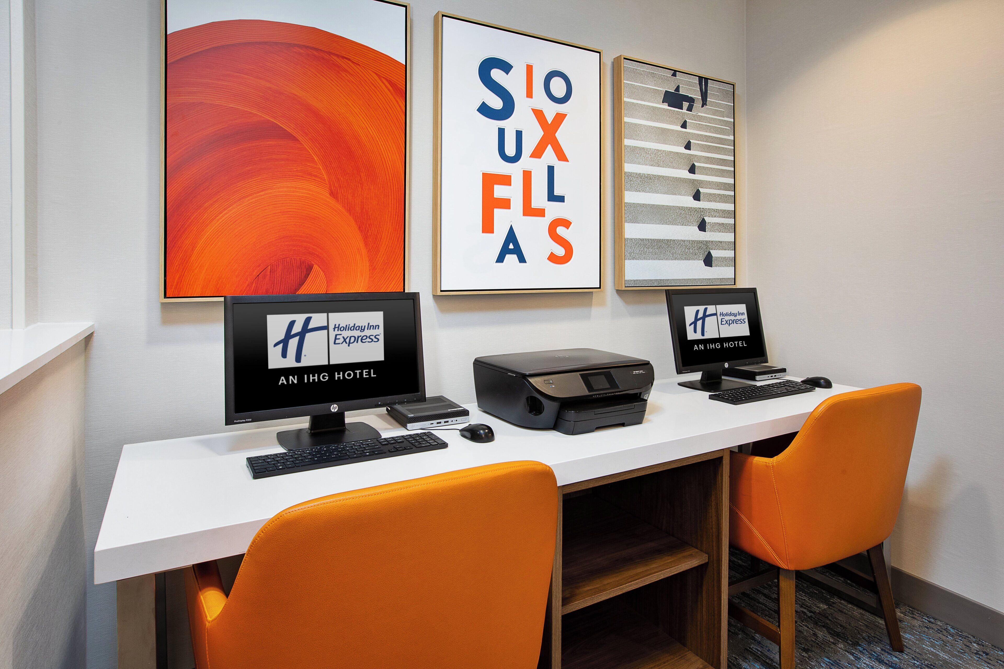 A hotel room with a desk, two computer monitors, a printer, and two orange chairs.