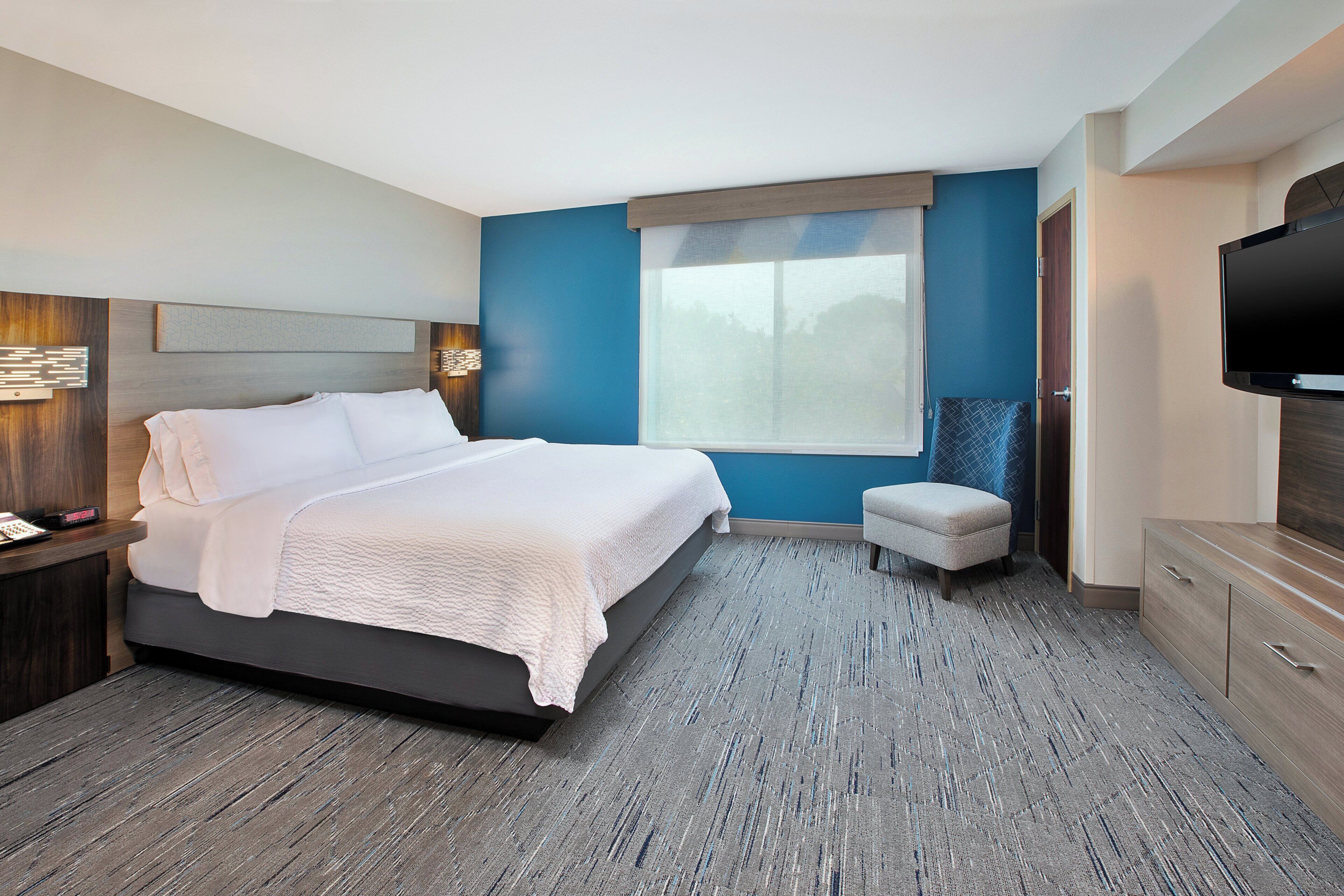 A modern hotel room with a large bed, a television, a small seating area, and a window with a view.