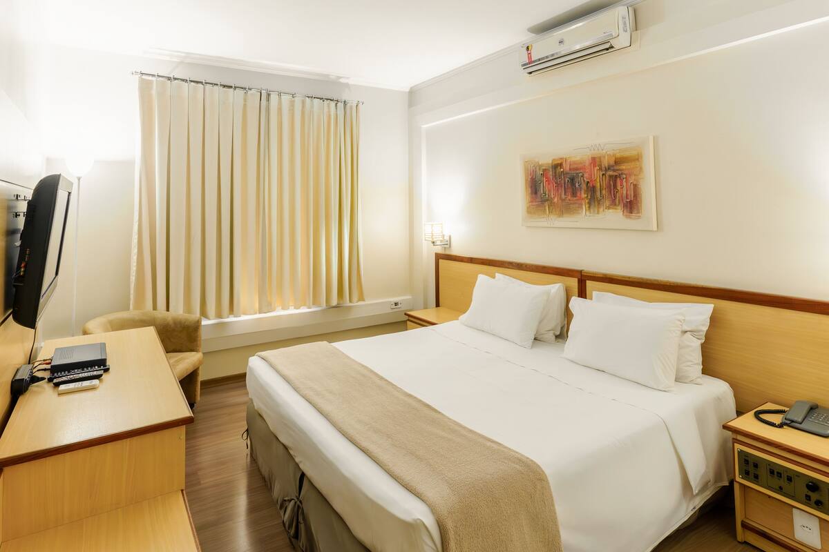 A hotel room with a bed, bedside table, desk, chair, and a window with curtains.