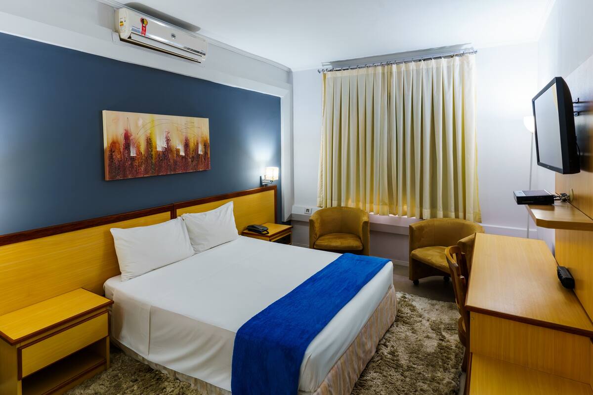 A hotel room with a bed, bedside tables, a desk, a chair, a television, a painting on the wall, and a window with curtains.