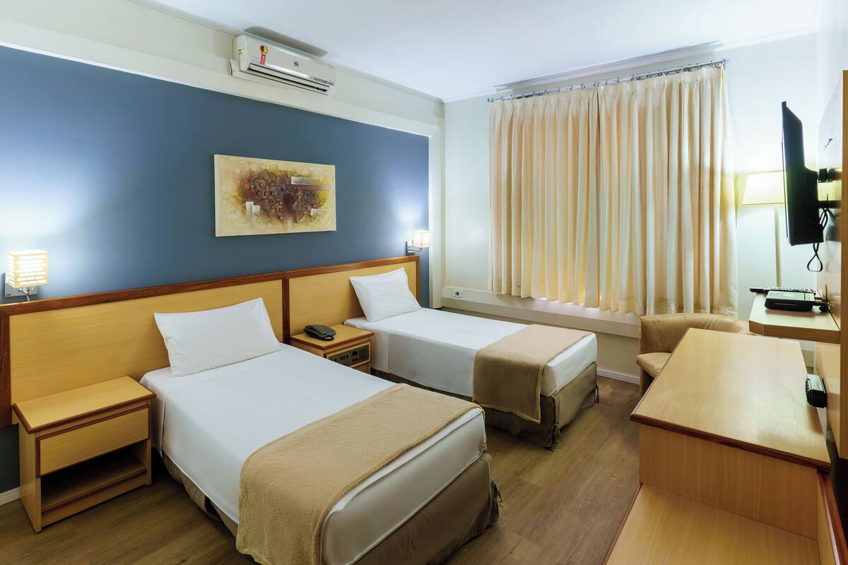 A hotel room with two beds, a desk, a chair, a TV, and a window with curtains.