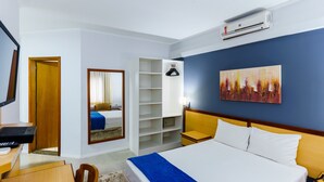 A hotel room with a bed, a desk, a chair, a wardrobe, a painting, and an air conditioning unit. - Hotel Augusta Boulevard (São Paulo)