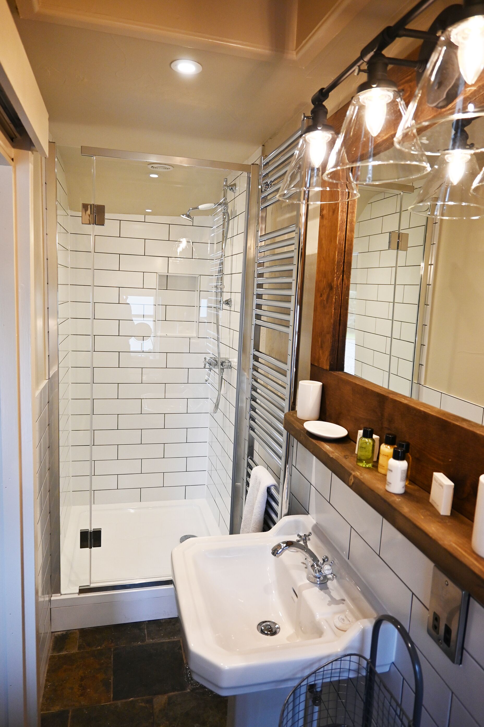 A bathroom with a glass-enclosed shower, white tiled walls, a wooden vanity with a mirror, and a white sink.