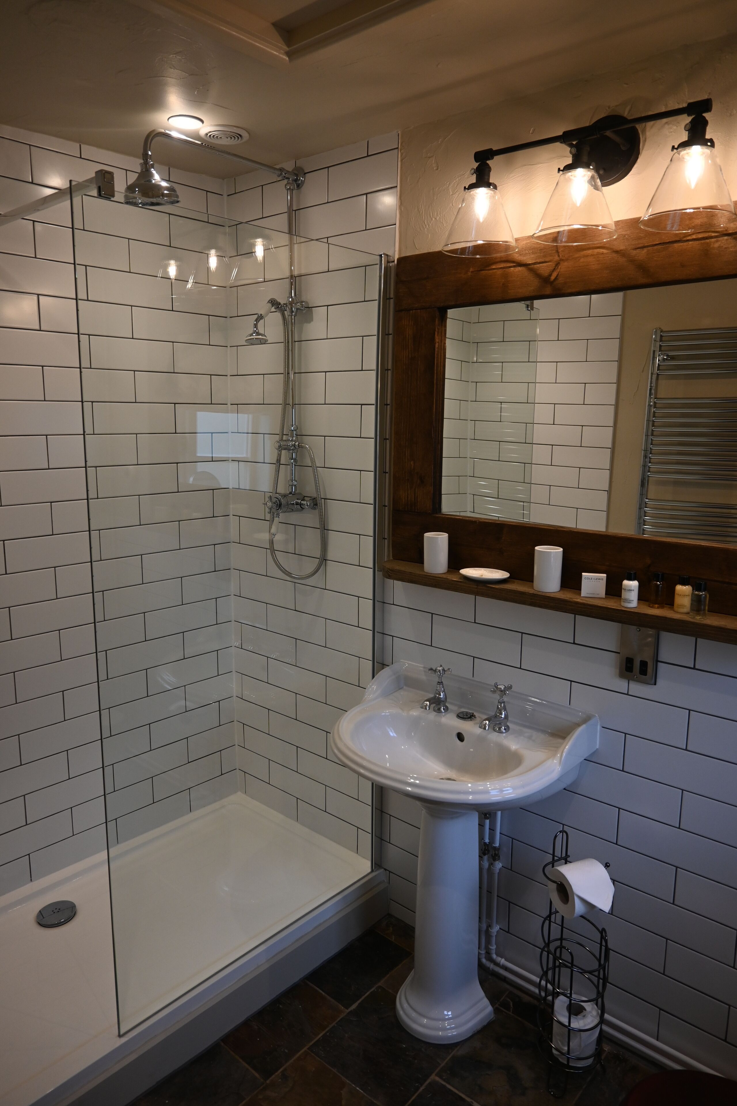A bathroom with a glass-enclosed shower, a pedestal sink, and a mirror with a wooden shelf above it.