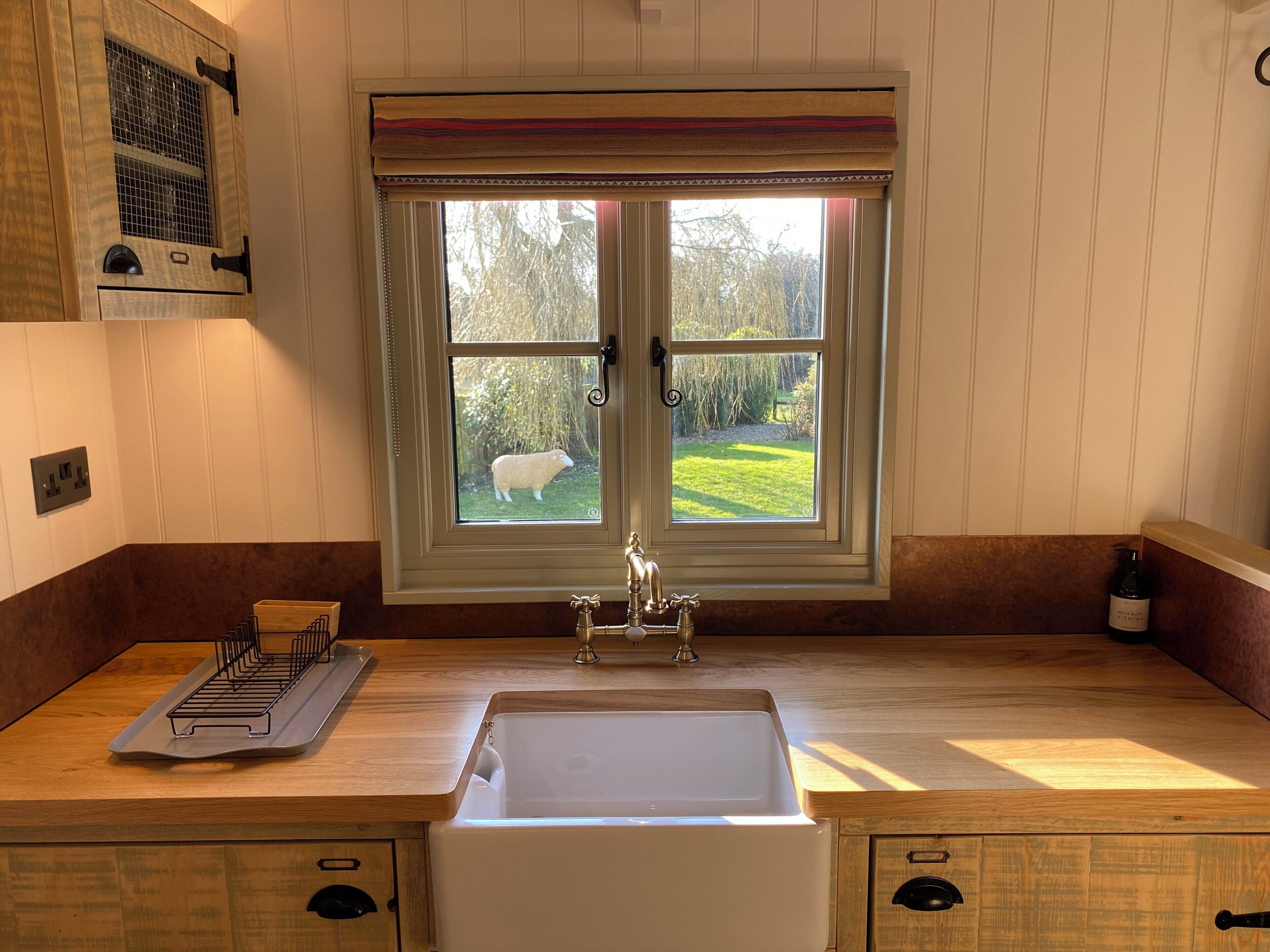 A kitchen with a double sink, wooden countertops, a window with a view of a sheep, and a hanging light fixture.