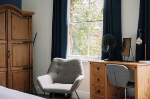 Una habitación con un armario de madera, un sillón gris, un escritorio con una silla, un ventilador de pie y una lámpara.