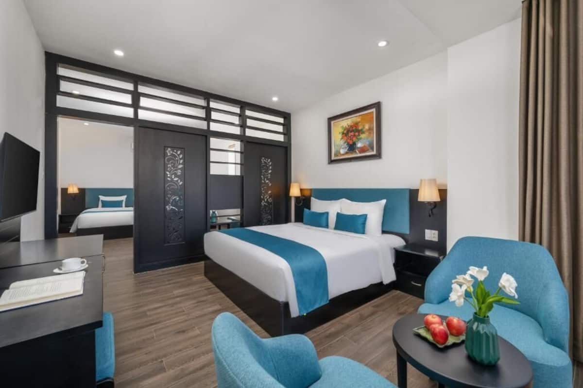 A modern hotel room with a large bed, a desk, a chair, a small table with flowers, and a painting on the wall.