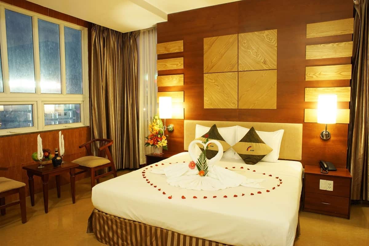 A hotel room with a large bed, two pillows, a nightstand, a chair, a table with a vase of flowers, and a window with curtains.