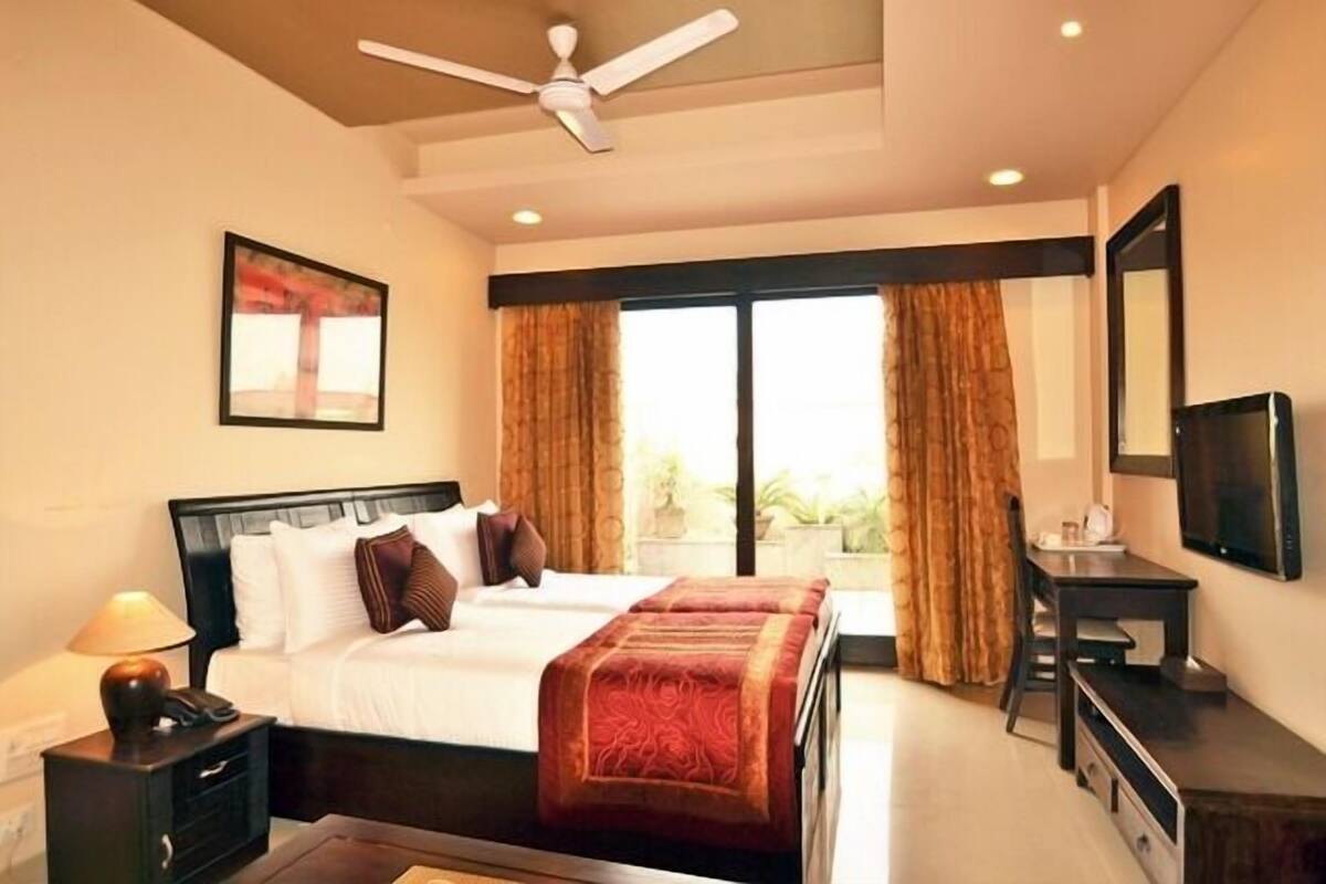 A hotel room with a bed, bedside table, lamp, TV, and a view of the outdoors.