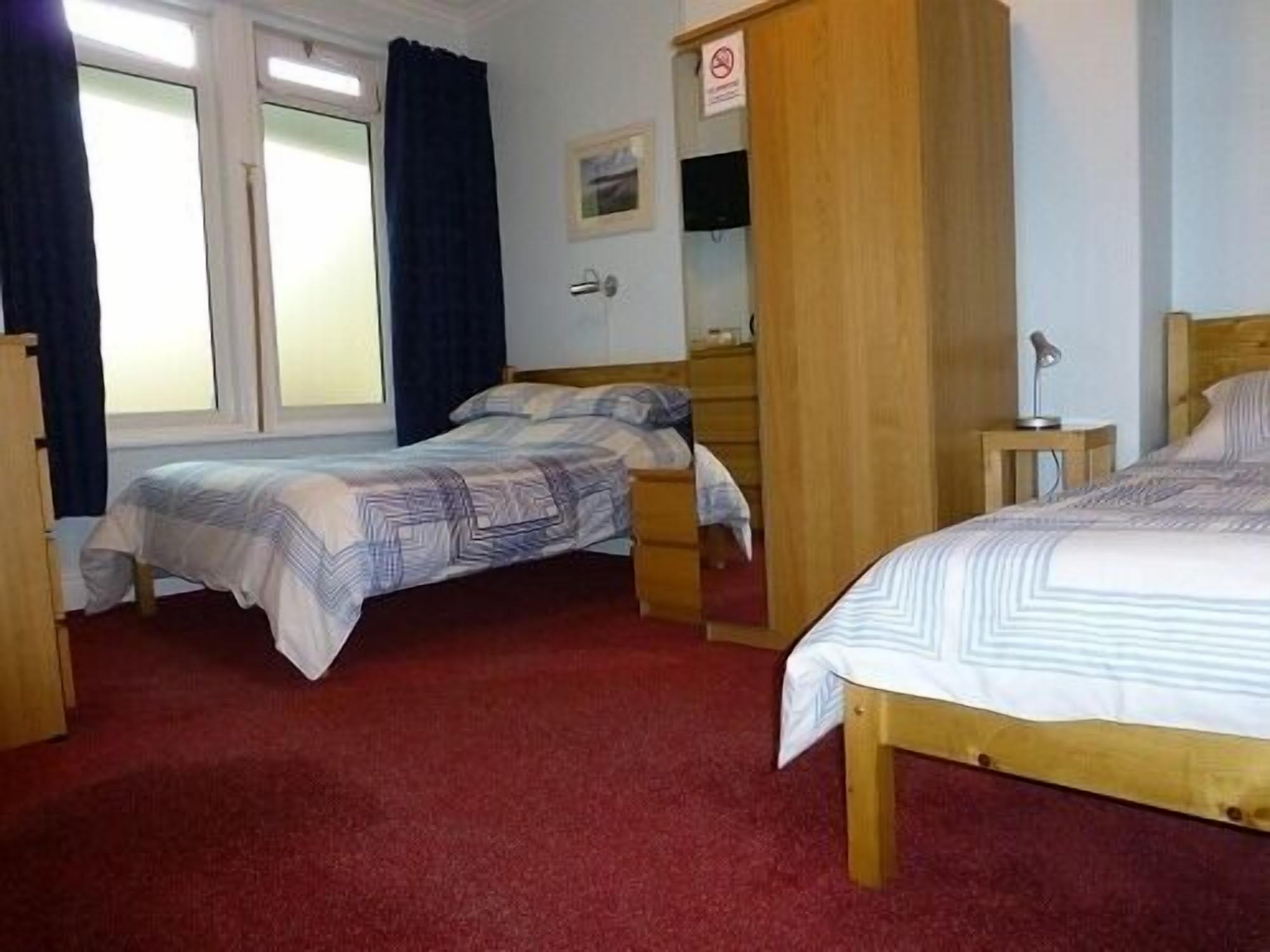A room with two single beds, a wooden wardrobe, a desk with a lamp, and a window with curtains.