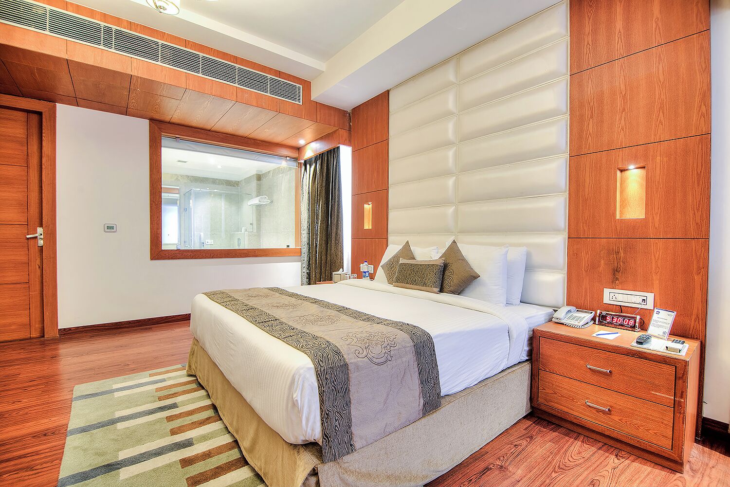A hotel room with a large bed, a wooden wardrobe, a bedside table with a phone and a clock, and a window with curtains.