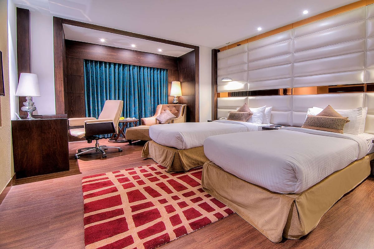 Quarto de hotel com duas camas, uma escrivaninha, uma cadeira, um abajur e um tapete vermelho estampado.