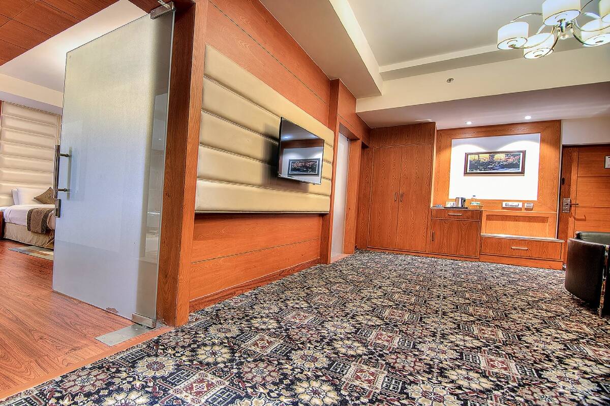 Quarto de hotel com piso de madeira, carpete estampado, cama, televisão fixada na parede e mesa com cadeira.