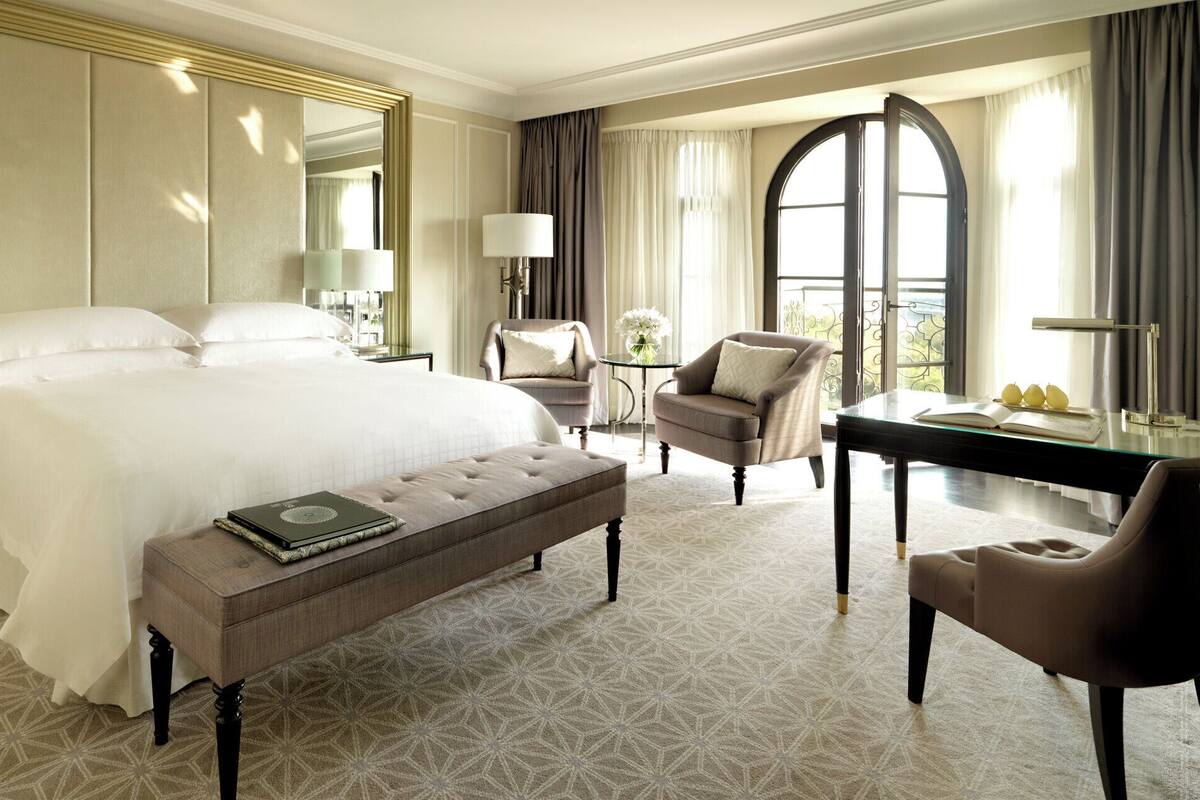 A hotel room with a large bed, a desk, a chair, and a bench.