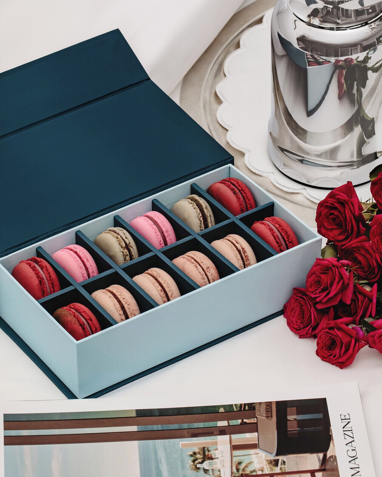Una caja de macarons con tapa celeste, sobre una superficie blanca junto a un candelabro plateado y rosas rojas.