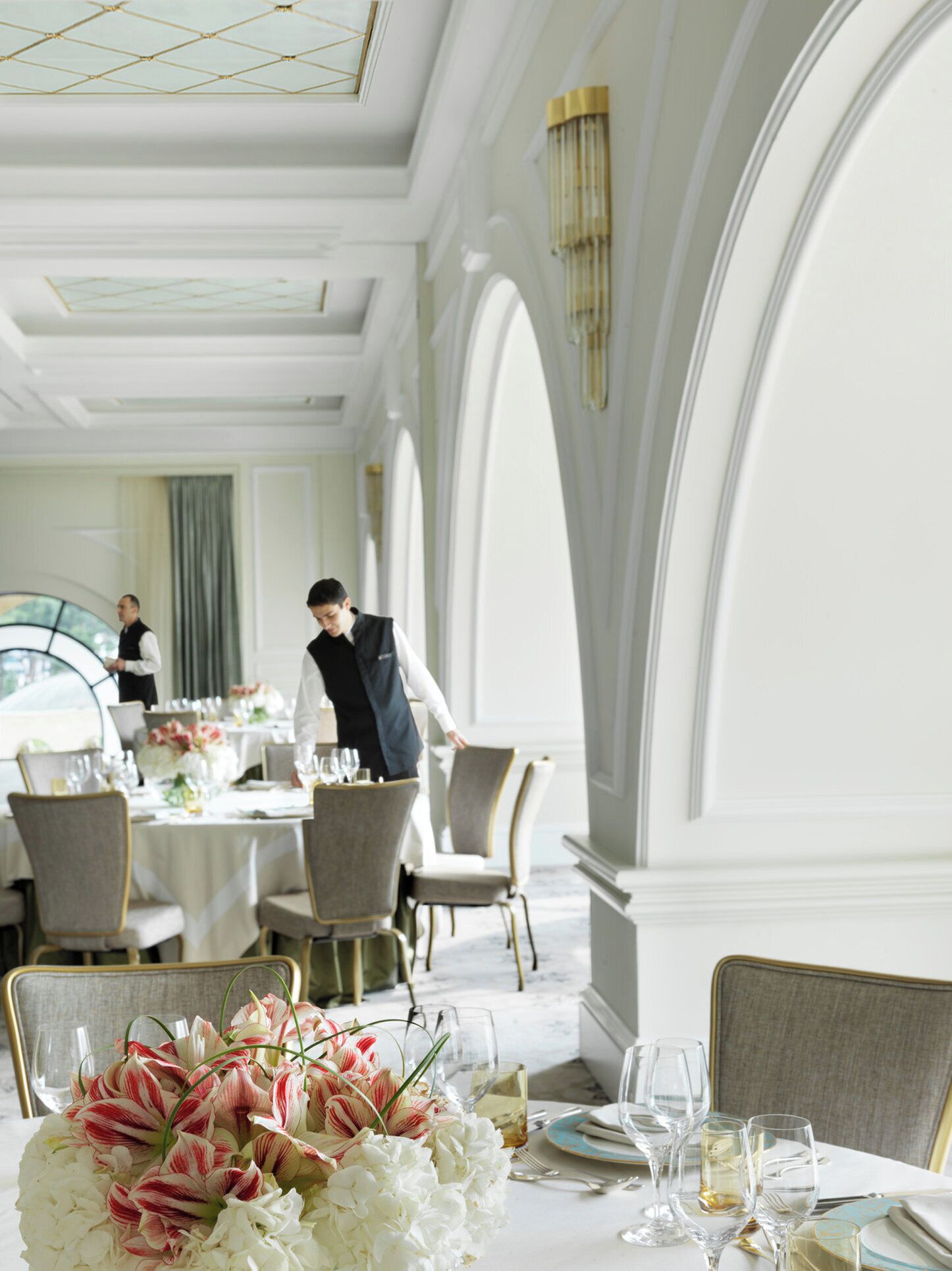 A formal dining room with tables set for service, elegant chairs, and a large floral centerpiece.