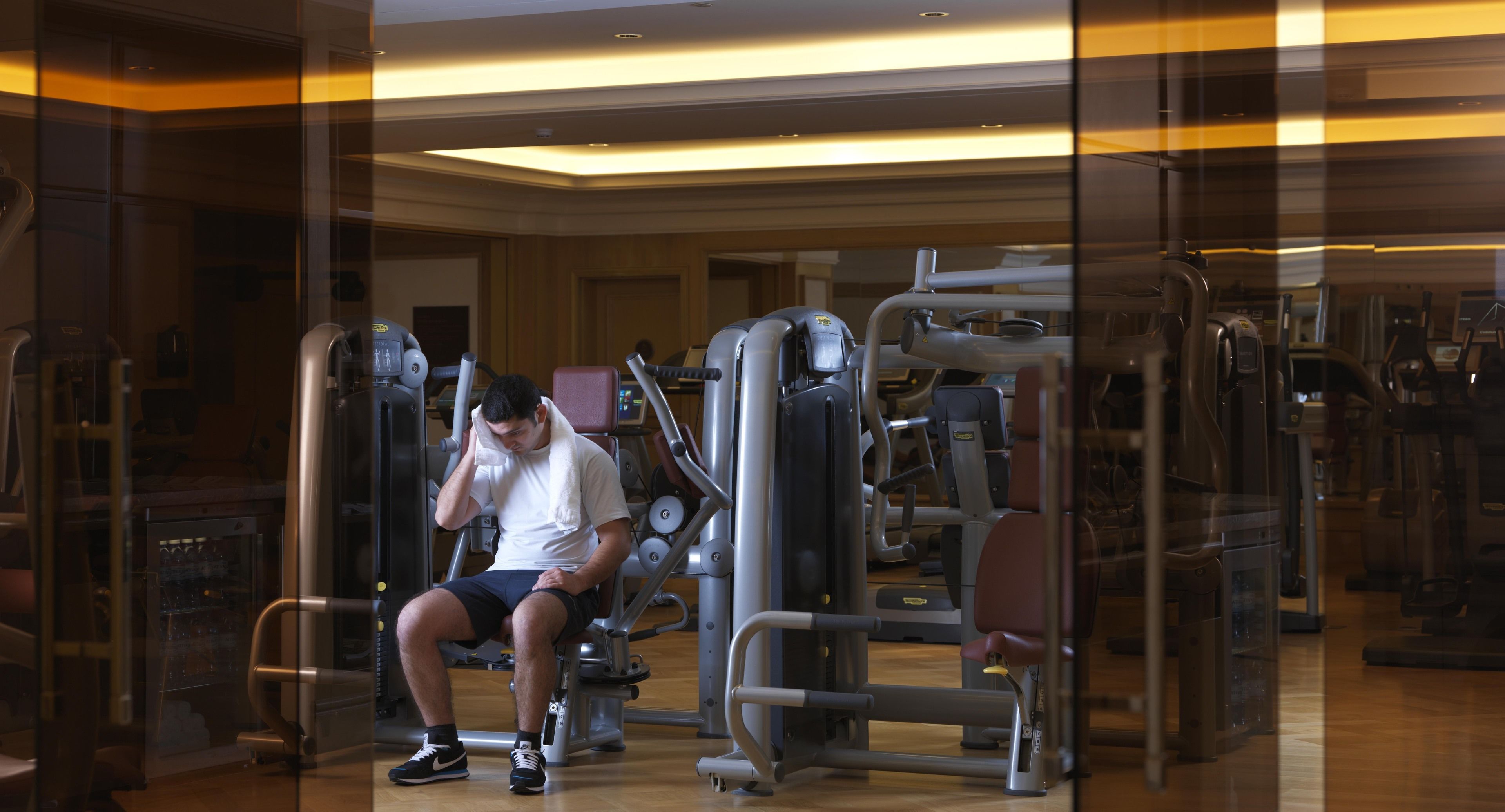 Ein Fitnessstudio mit verschiedenen Trainingsgeräten, eine Person sitzt auf einer Bank, und eine Glasschrankwand spiegelt das Innere wider.