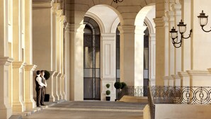 A grand hallway with classical architecture, featuring columns, arches, and a chandelier.