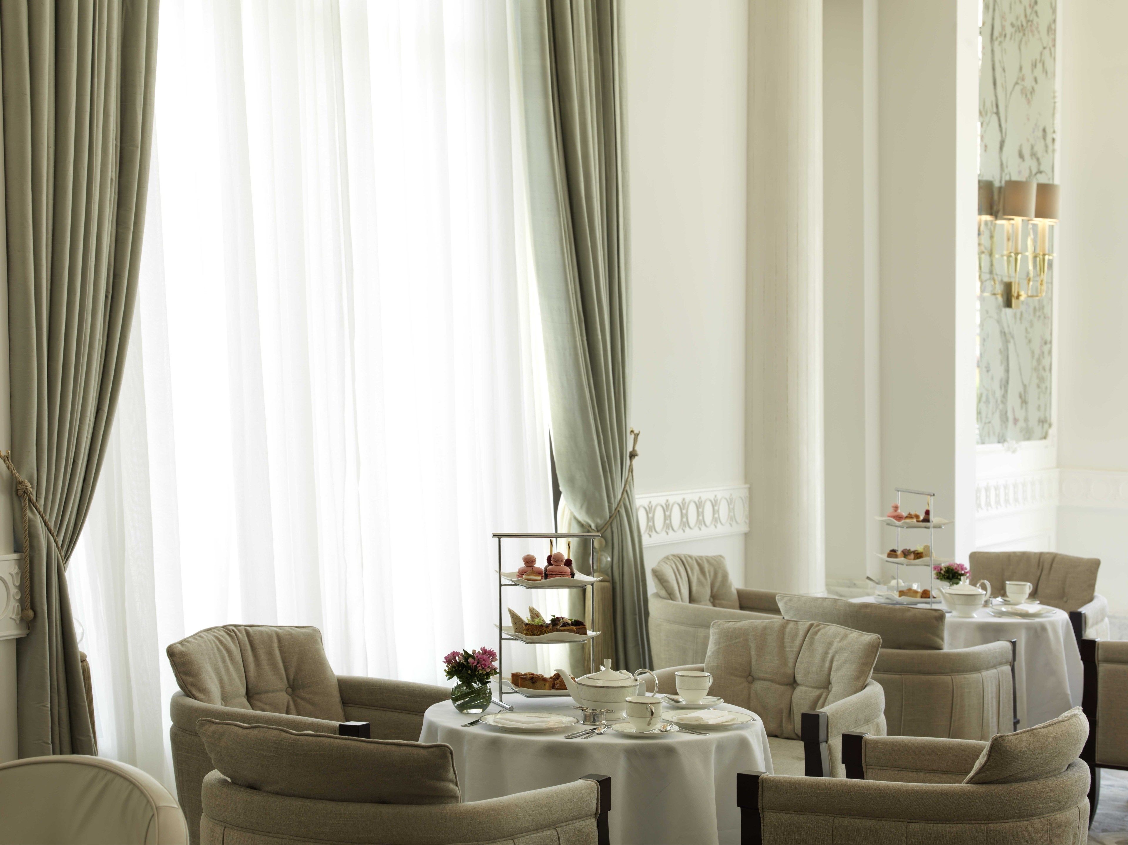 A formal dining room with multiple tables and chairs, a tiered dessert display, and large windows with sheer curtains.