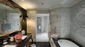 Une salle de bain luxueuse avec un grand miroir, des murs en marbre, une baignoire et une commode agrémentée d'un vase de fleurs.