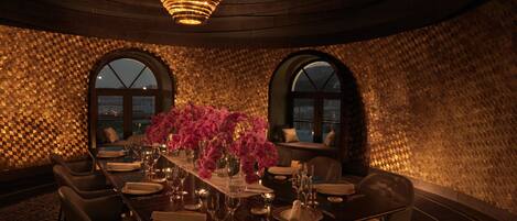 A dining area with a long table set for six, surrounded by chairs, with a chandelier above and a patterned wall behind.