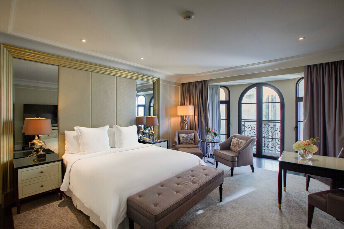 A hotel room with a large bed, a sitting area with a table and chairs, a mirror, and a view of the city through large windows.