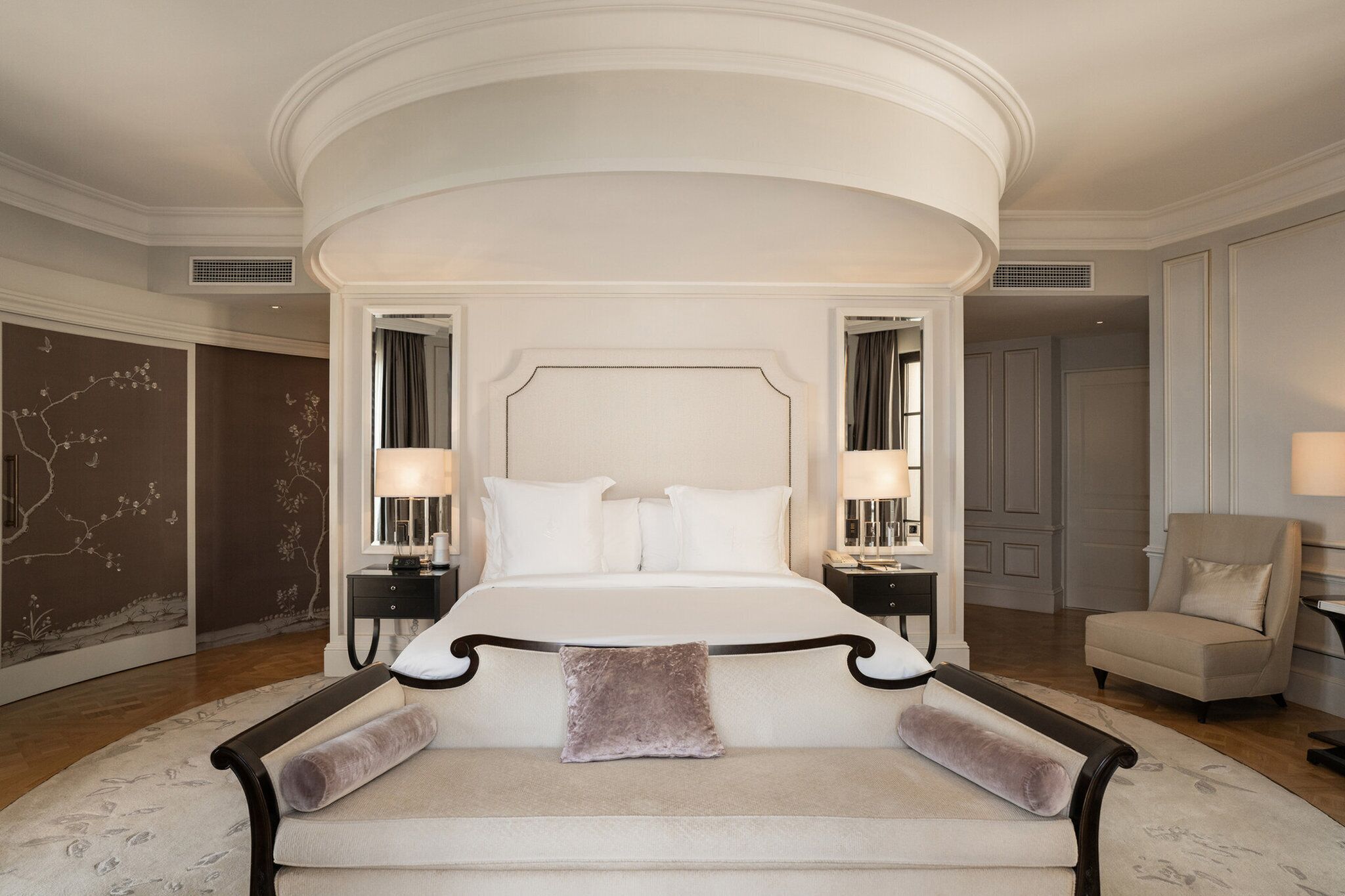 A four-poster bed with a white headboard, two bedside tables with lamps, a beige armchair, and a patterned rug.