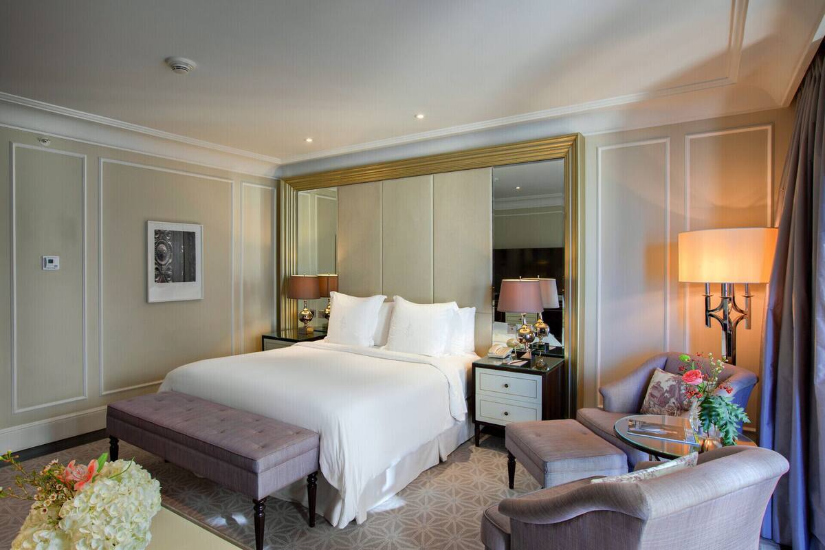 A hotel room with a large bed, a bench, a chair, a nightstand, a lamp, and a mirror.
