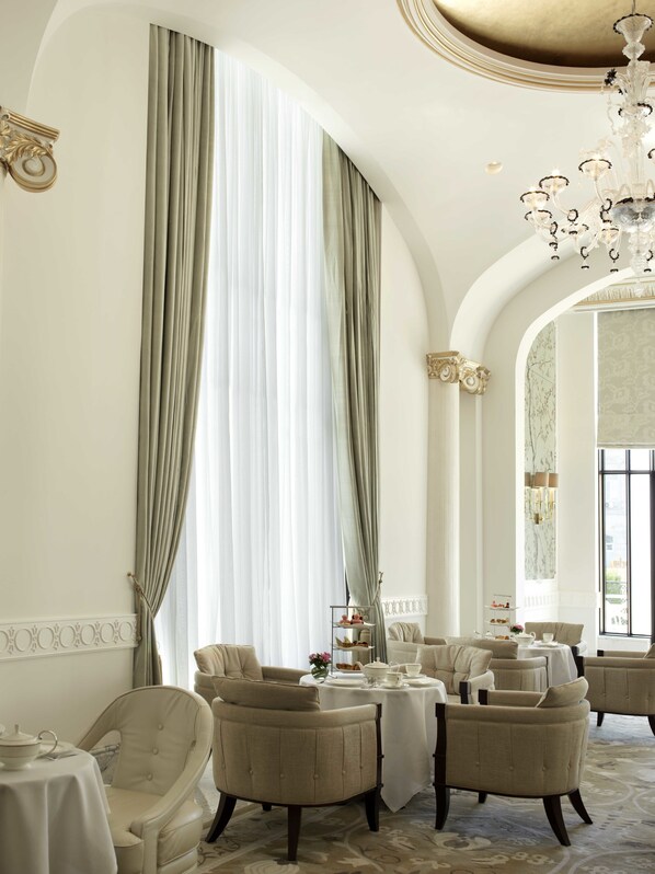 A room with a high arched ceiling, large windows with sheer curtains, and a chandelier. There are multiple seating areas with tables set for dining.