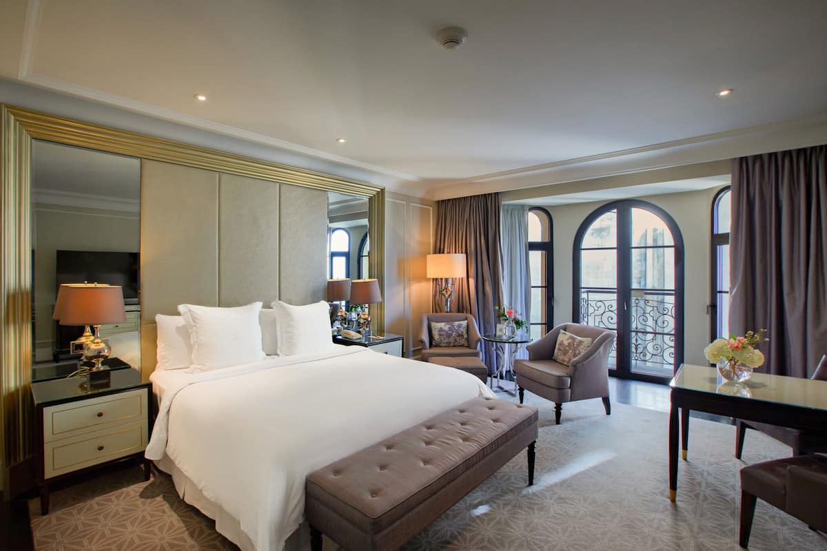 A hotel room with a large bed, a sitting area with a table and chairs, a mirror, and a view through the window.