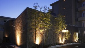 Un bâtiment moderne doté d’une façade en pierre et de fenêtres illuminées, entouré d’un espace paysager agrémenté d’arbres.