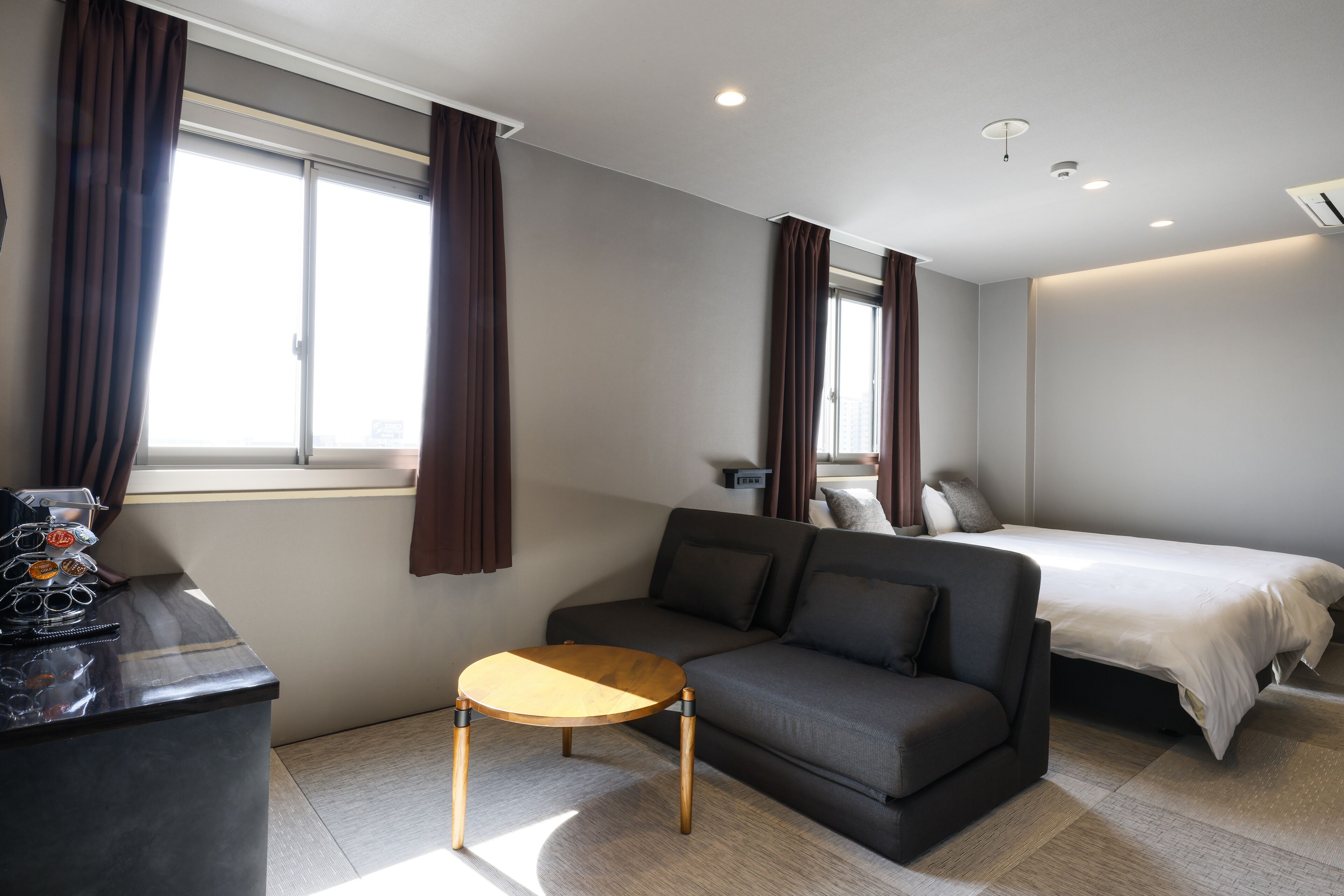 Una habitación de hotel moderna con sofá, una mesita redonda, cama y escritorio con computadora y lentes de sol.