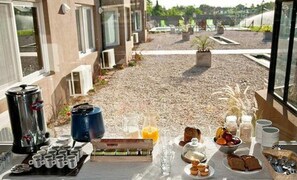Desayuno con café, té, pastelería y fruta en un balcón con vistas a un área de grava, edificios y vegetación.