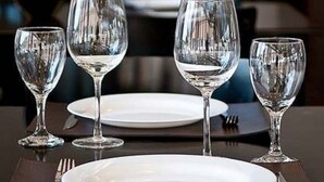 Mesa de comedor preparada con cuatro copas de vino y platos, con un fondo desenfocado que sugiere un restaurante.