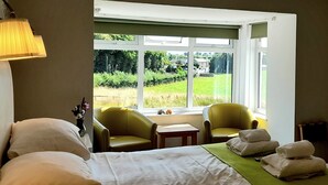 Habitación de hotel con cama, dos sillones, una mesita y vistas a un paisaje verde a través de amplios ventanales.
