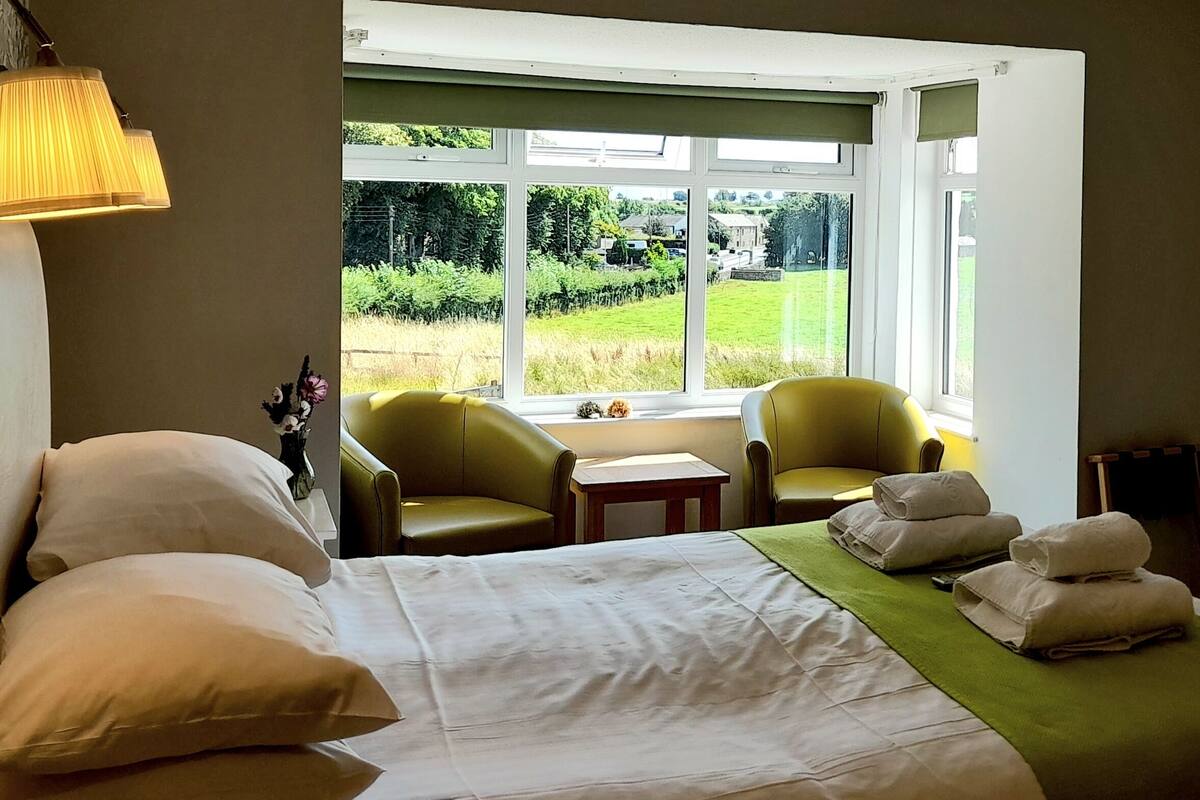 A hotel room with a bed, two armchairs, a small table, and a view of a green landscape through large windows.