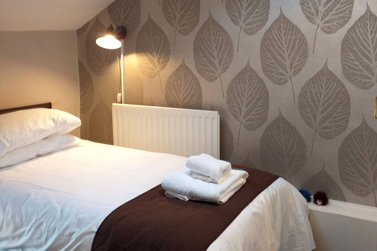 A hotel room with a bed, a radiator, a wall with a leaf pattern, and a nightstand with a lamp.