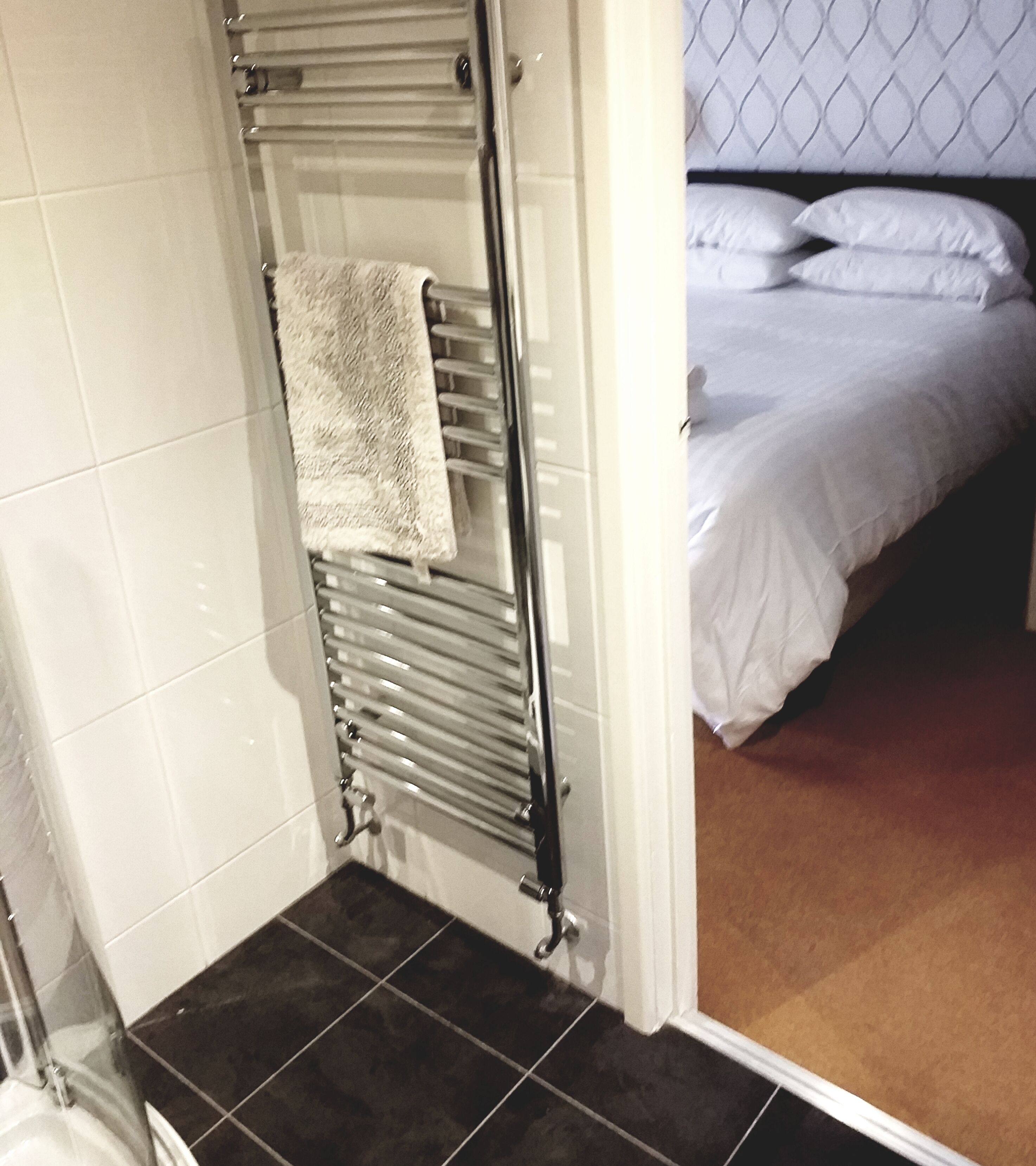 A bathroom with a towel rack, a shower area, and a bed with white bedding.