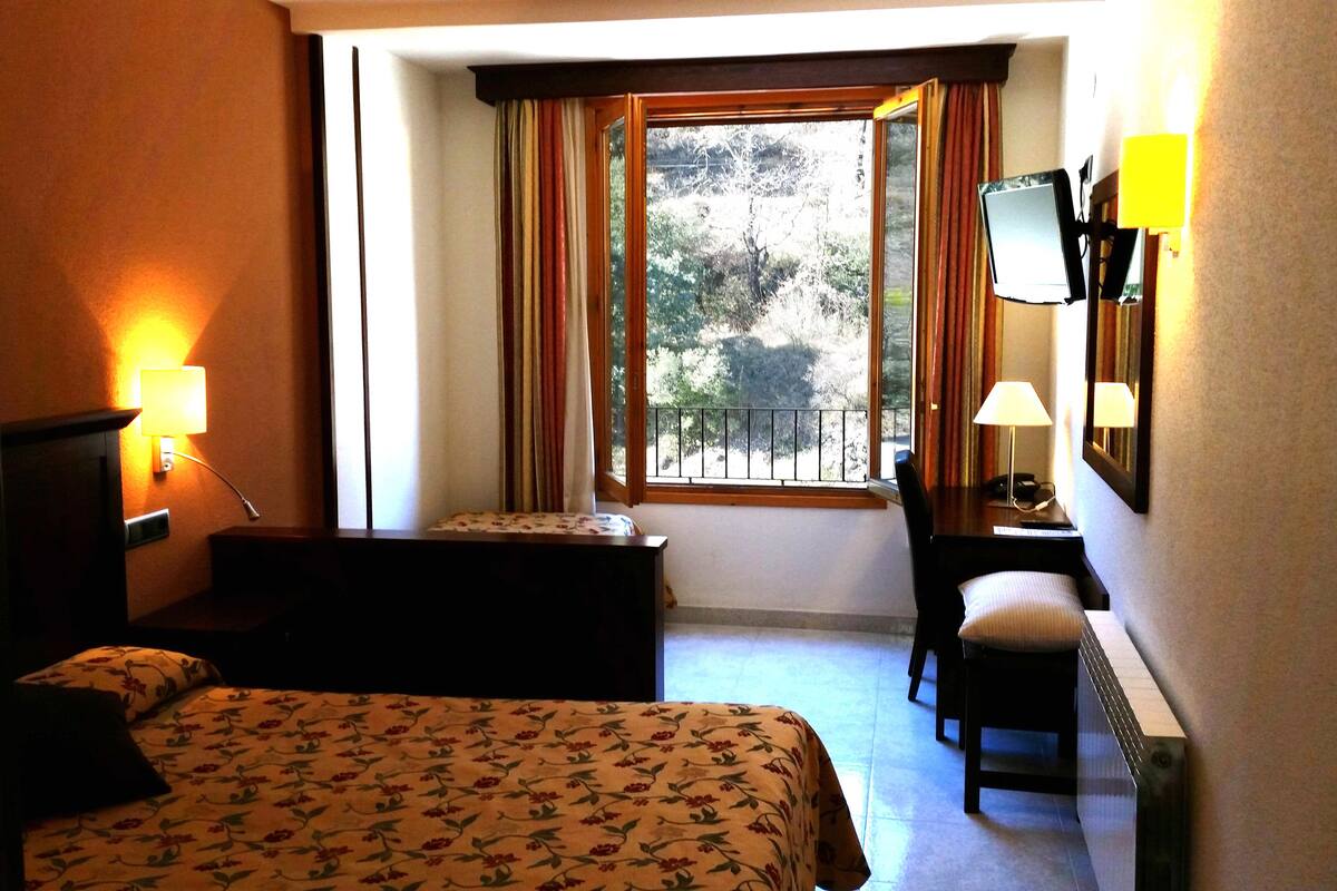 A hotel room with a bed, a desk, a chair, a window with curtains, a TV, and a lamp.
