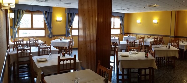 Um restaurante com várias mesas e cadeiras, painéis de madeira e janelas grandes com cortinas.