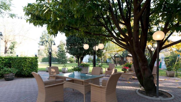 An outdoor patio with wicker furniture and a glass table, surrounded by trees and a hedge. - Hotel Ristorante ai Campi Di Marcello (Monfalcone)