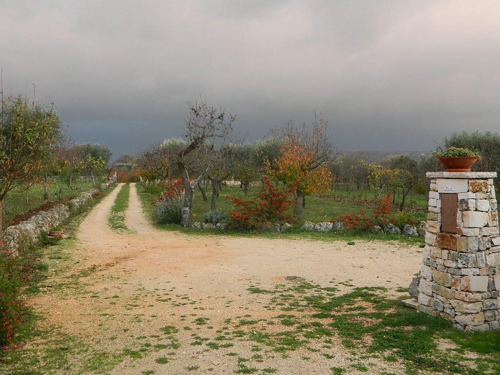 Uma trilha de terra serpenteia por um jardim com muros de pedra e uma estrutura de pedra com um vaso em cima.