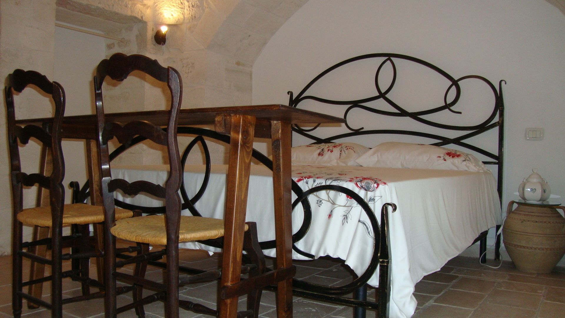 Une chambre à coucher comprenant un lit, deux chaises en bois, une tête de lit en métal, un couvre-lit blanc et un petit pot décoratif.
