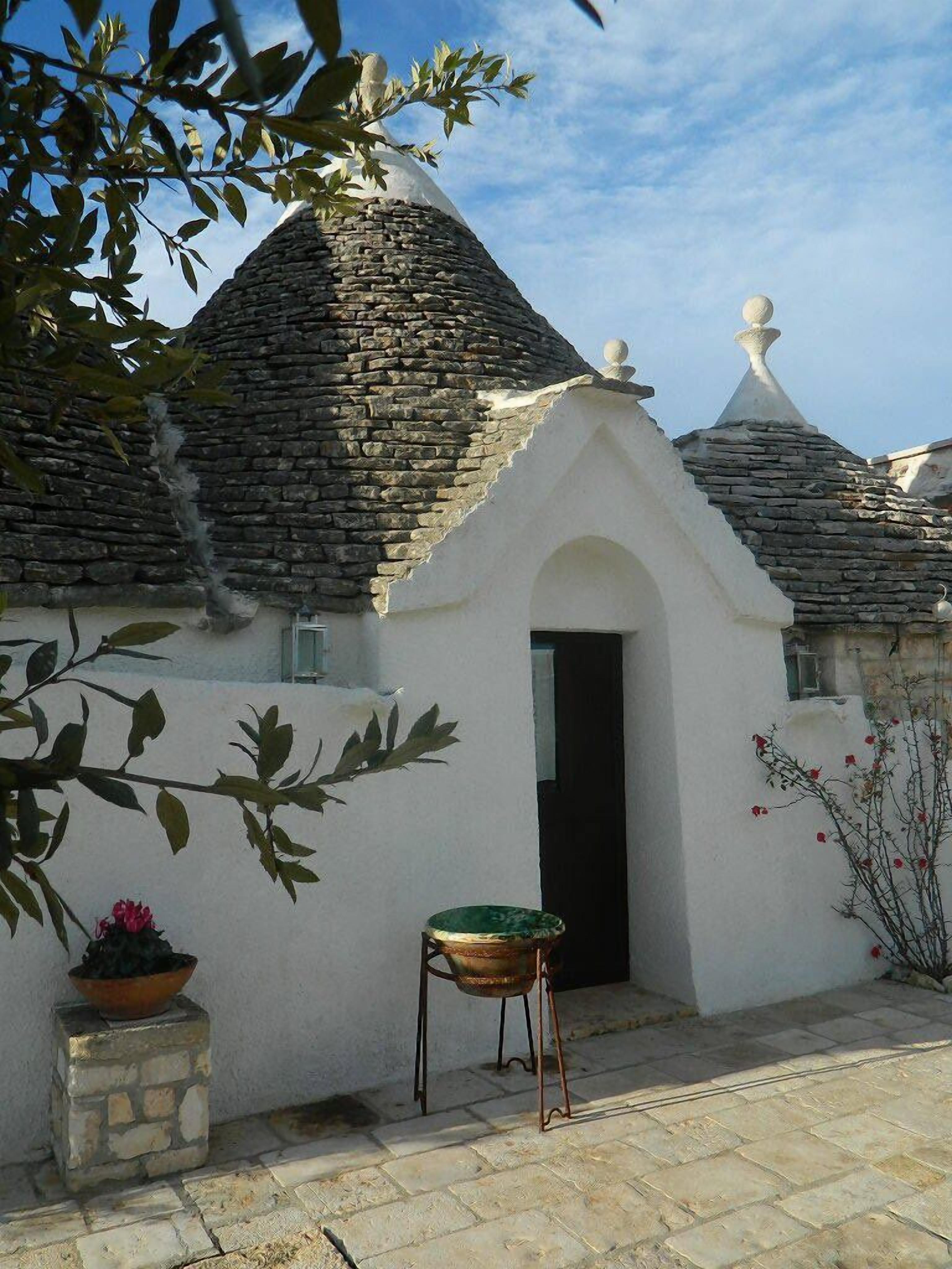 Un bâtiment traditionnel en pierre, doté d’une façade blanche, d’une porte noire et d’un petit jardin agrémenté d’une chaise en métal et de plantes en pot.