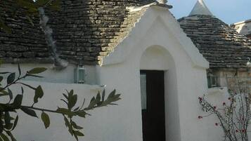 Un bâtiment traditionnel en pierre, doté d’une façade blanche, d’une porte noire et d’un petit jardin agrémenté d’une chaise en métal et de plantes en pot.