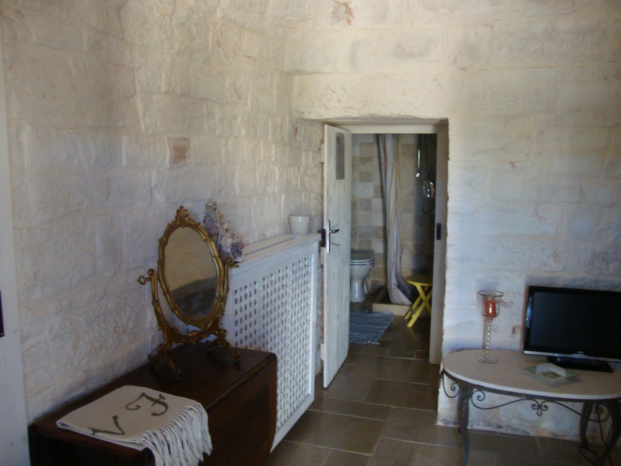 A room with a stone wall, a wooden dresser with a mirror, a television on a small table, and a door leading to a bathroom.