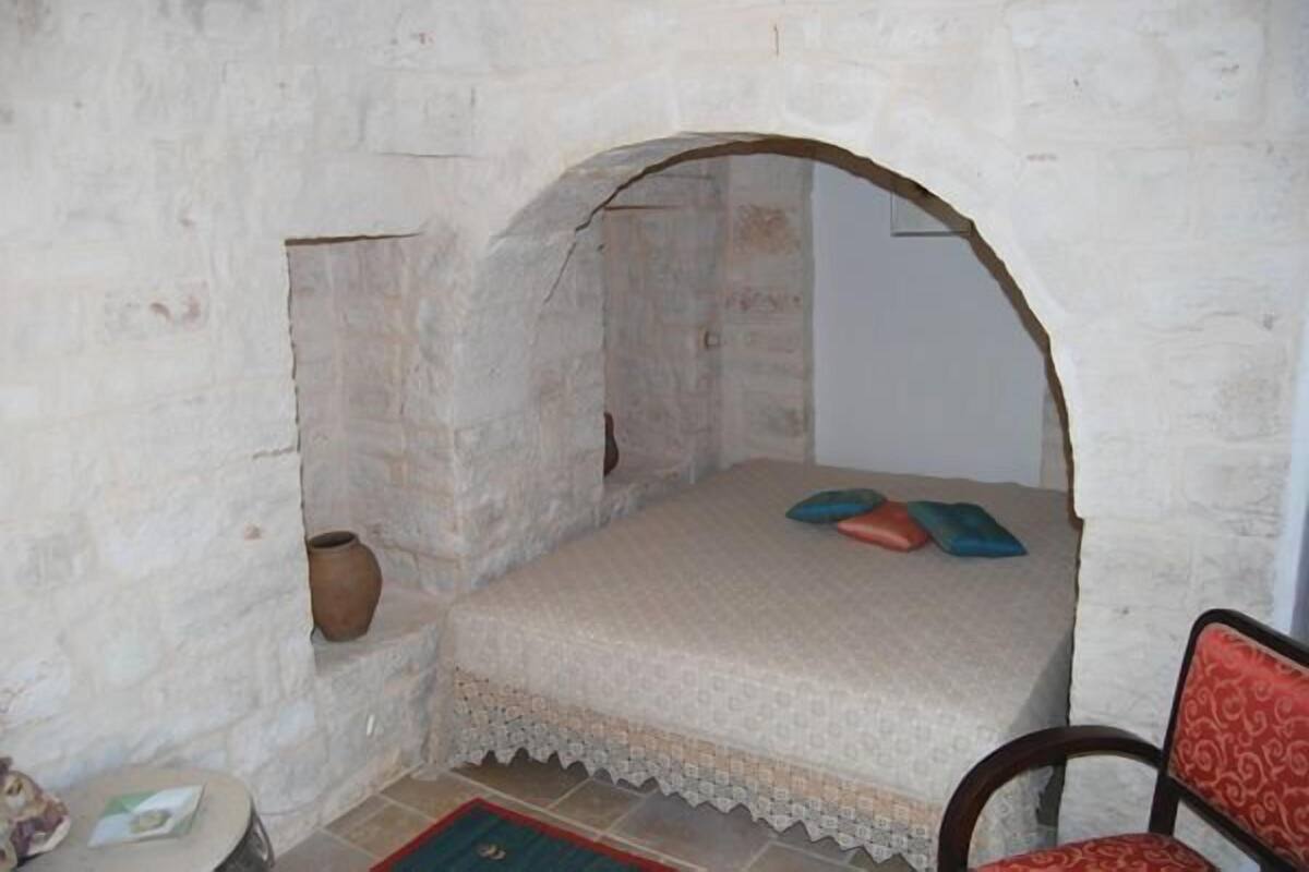 Pequeña habitación rústica con pared de piedra, una cama individual con colcha estampada, una mesita con un libro y una silla con cojín rojo.
