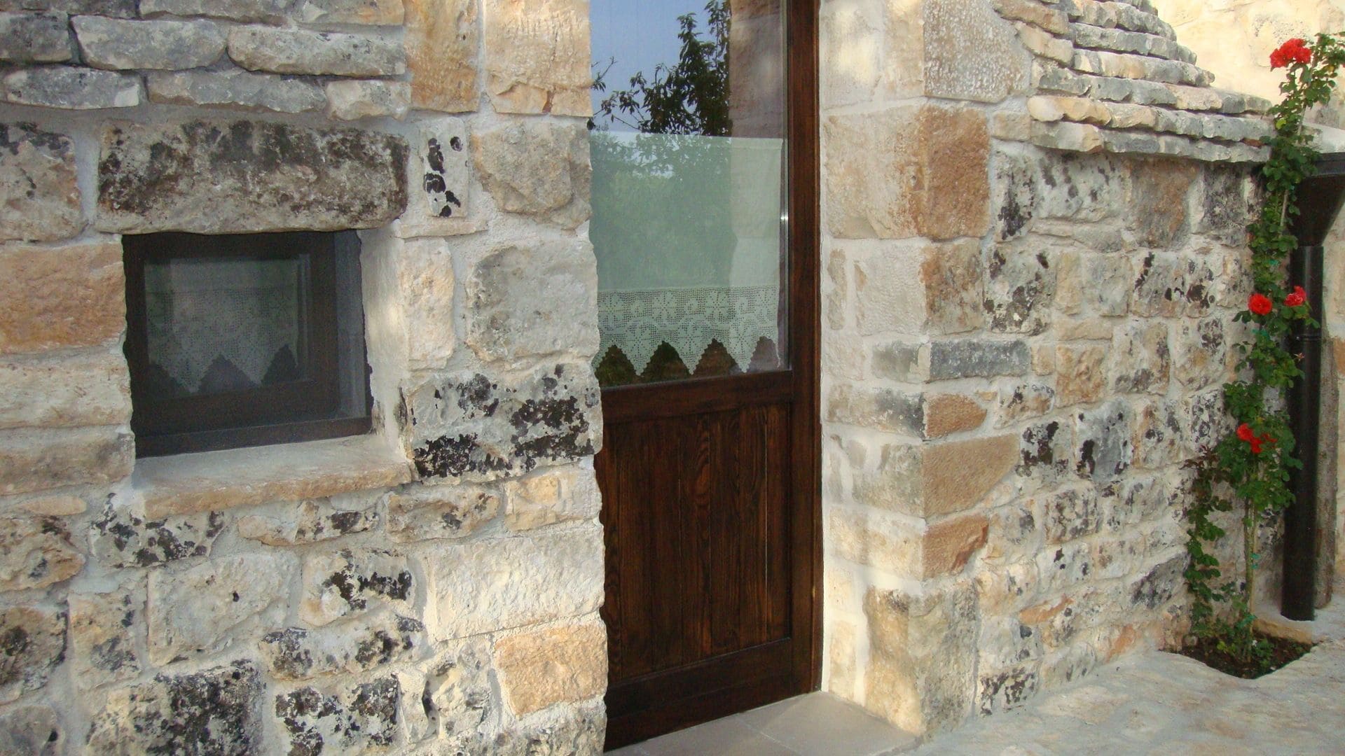 Un bâtiment en pierre avec une porte et une fenêtre en bois, un petit balcon agrémenté d’une plante, et un mur en pierre orné d’un motif décoratif.