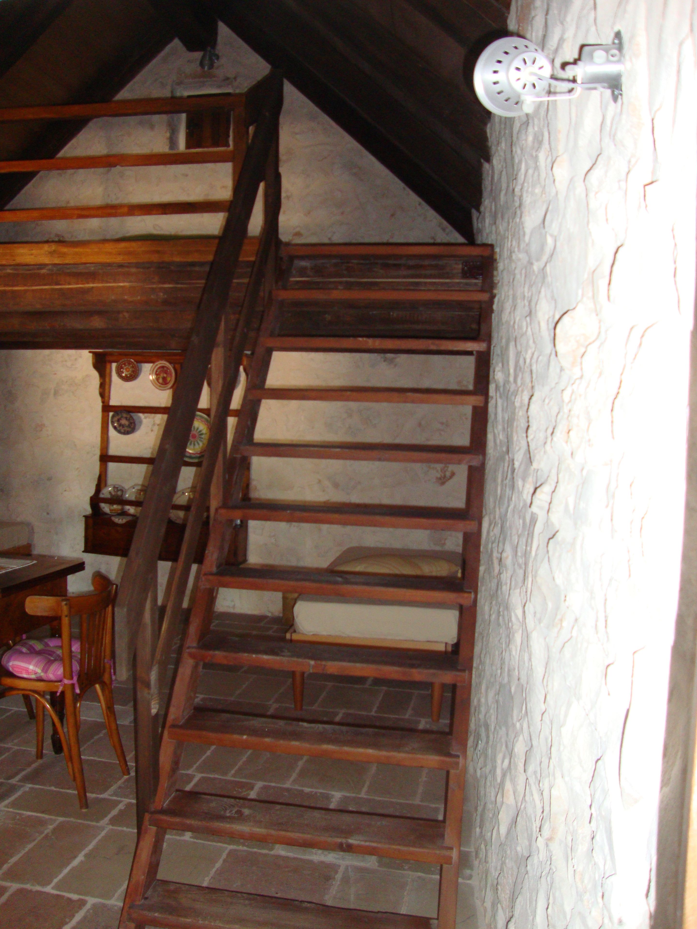 Un escalier en bois dans une pièce au style rustique, avec un sol en briques, une chaise en bois et un luminaire fixé au mur.