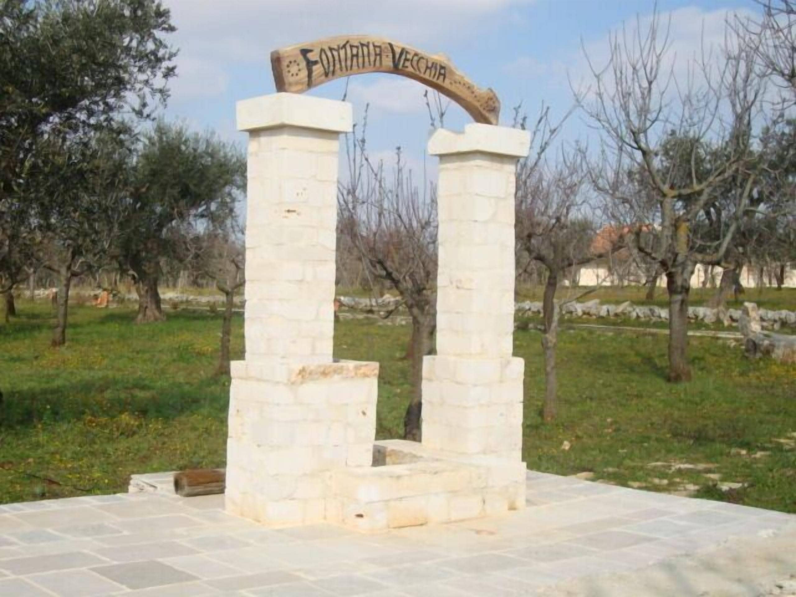 A stone monument with a wooden sign reading "Fontana Vecchia" in a grassy area with trees.